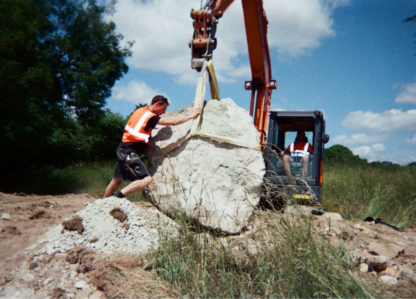 A construction worker in an orange safety vest and cap pushes a large chalk boulder being lifted by an excavator. The boulder is secured with straps attached to the excavator’s arm. The scene is set outdoors on a sunny day, with green trees and grass in the background.