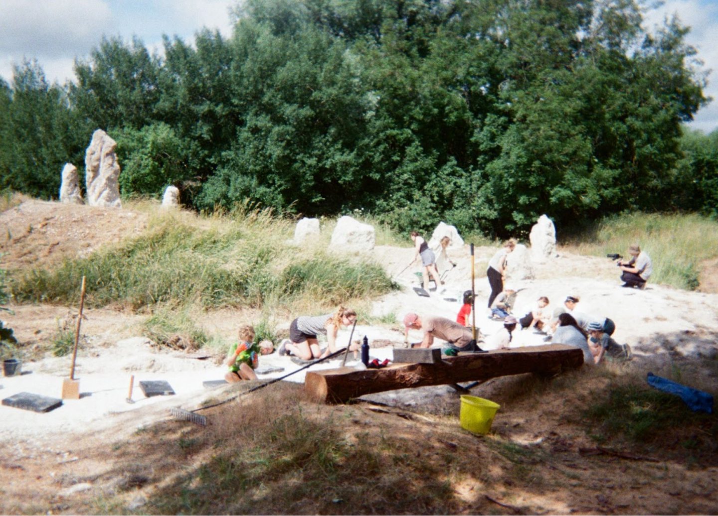 A group of adults and children participate in an outdoor chalk packing day in a grassy area. They are using tools to work on the ground, surrounded by tall trees and large standing stones. Equipment such as a yellow bucket, wooden beams, and black mats are scattered around. The sky is partly cloudy.