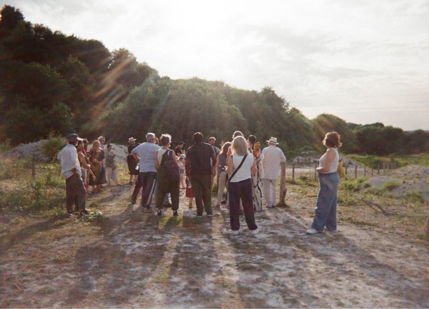 A group of people gathered outdoors on a chalky and grassy area surrounded by trees and bushes. The sun shines brightly, casting long shadows across the ground. Some individuals are standing, while others appear to be conversing or observing something. The scene has a peaceful, natural ambiance with sunlight filtering through the trees.