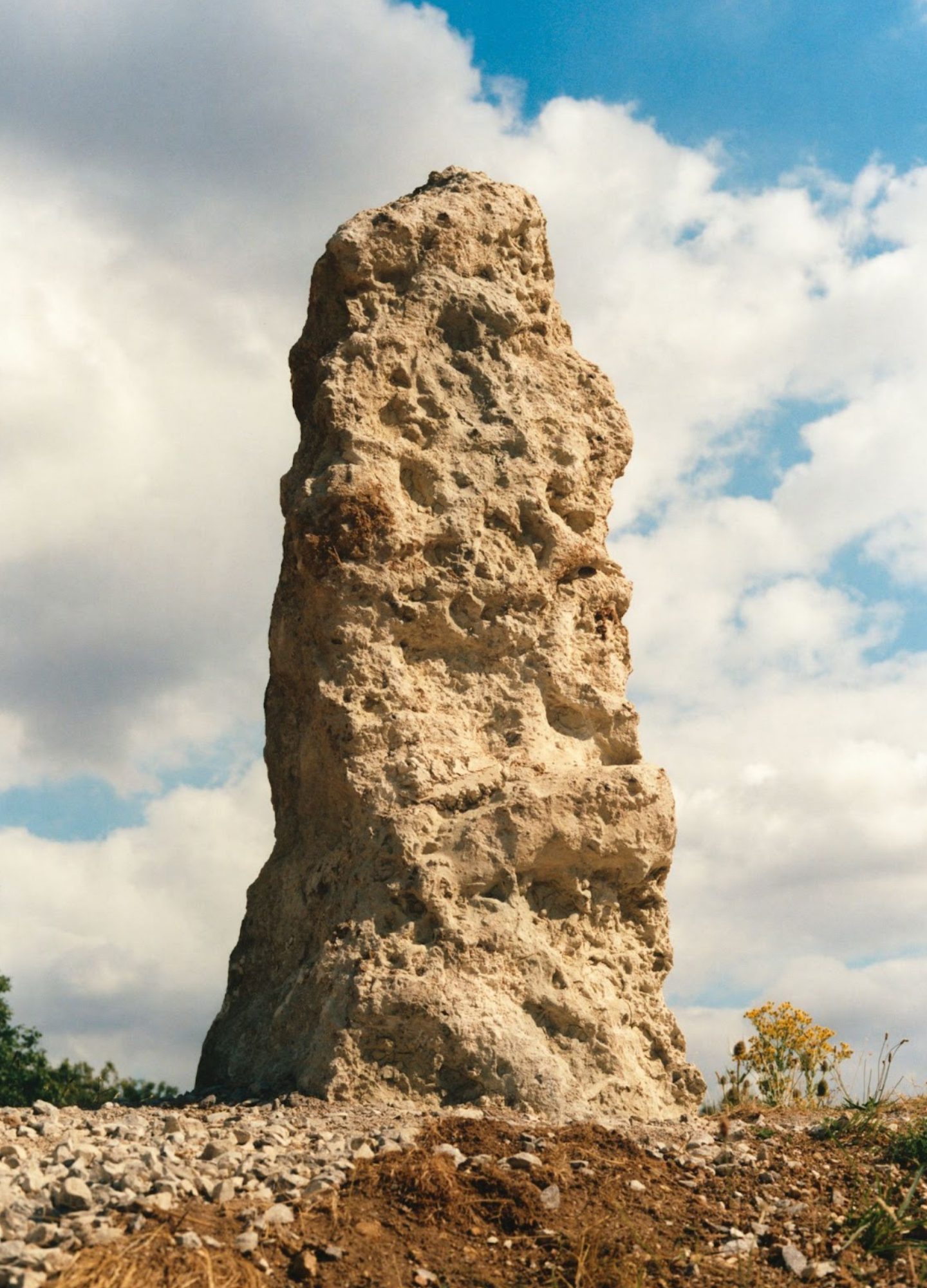 A tall, irregularly shaped rock formation (chalk) with a rough, textured surface stands upright against a partly cloudy sky. The ground around it is covered in small stones and chalk, with sparse vegetation visible to the right.