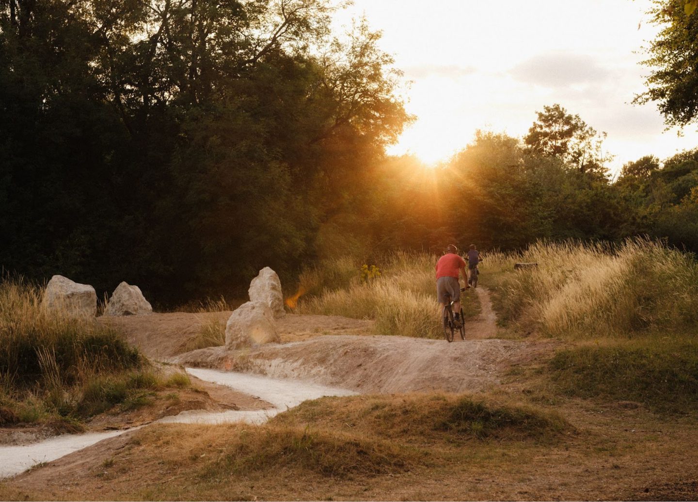 A person in a red shirt and grey shorts rides a bicycle along a chalk path through a grassy area with large rocks. The setting sun casts a warm glow over the scene, which is surrounded by trees and bushes, creating a peaceful, natural atmosphere.