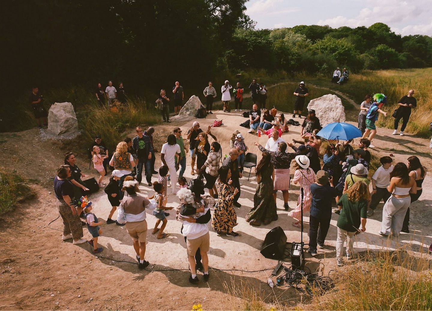 A large group of people gathered in a circular outdoor area surrounded by tall chalk, grass and trees. Some are dancing, others are standing or seated on a blanket on the ground. Children are playing and adults are socialising, creating a lively scene with a mix of casual and festive clothing.