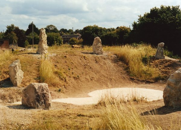 A grassy, slightly hilly landscape featuring several large chalk standing stones arranged in a circular formation. The ground has patches of dry grass and chalk. Trees and partially visible buildings appear in the background beneath a partly cloudy sky.