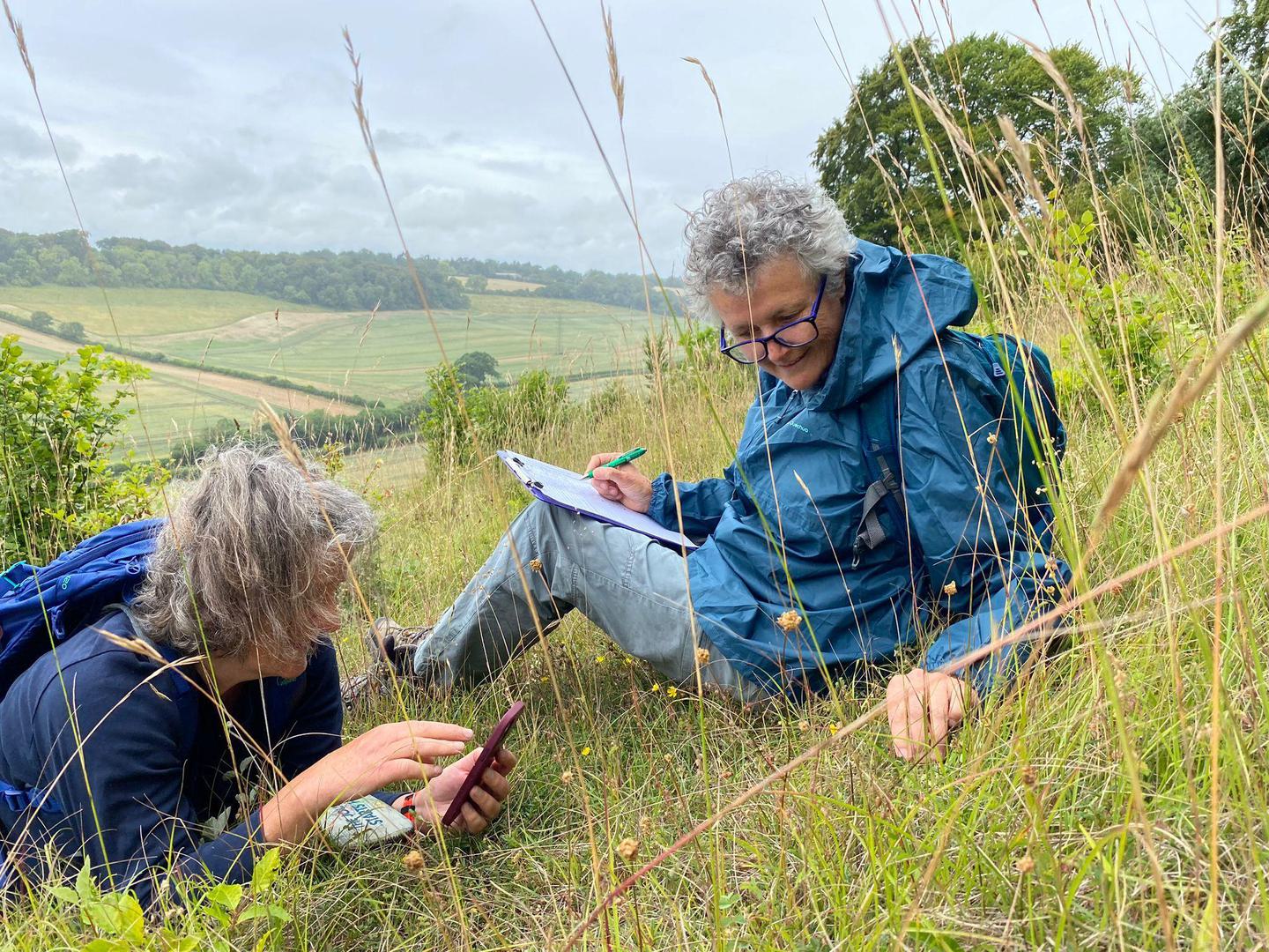 Two people in outdoor clothing lying on a grassy hillside, one using a phone and the other writing on a clipboard, with a scenic view of fields and trees in the background.