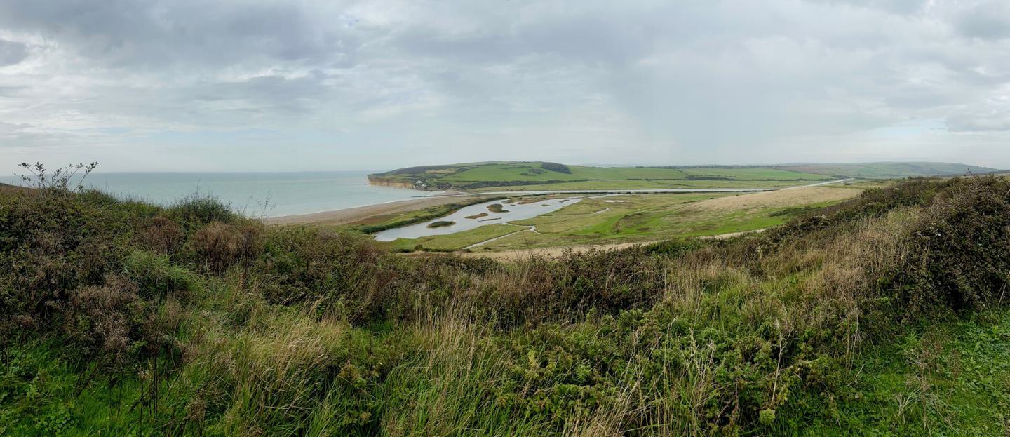 Panoramic view of the South Downs National Park with rolling green fields, cliffs, a sandy beach, and calm coastal waters under a cloudy sky.