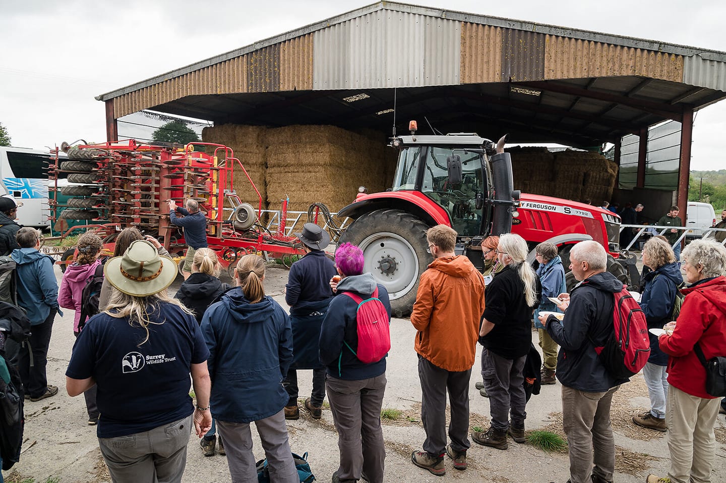 A group of people engages in conversation with a farmer on a National Trust farm within the Stonehenge Estate. The scene features a bright red tractor parked nearby, with bales of hay scattered around and a rustic barn visible in the background.