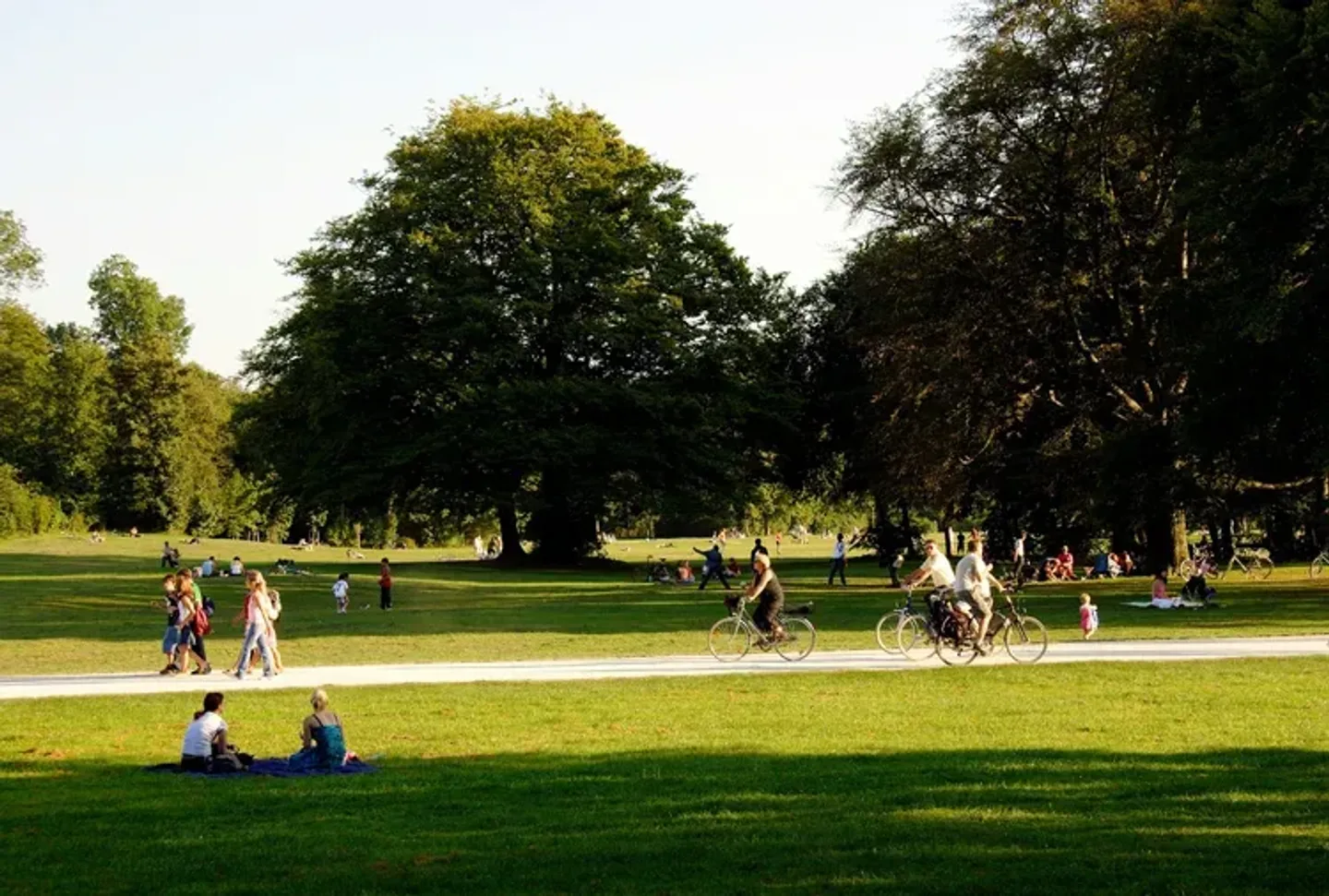 A sunny day in a park where people are enjoying various activities. In the foreground, two adults and a child sit on a blanket on the grass. Nearby, others walk along a paved path or ride bicycles. Large trees provide shade in the background, with more people relaxing on the grass under a clear, cloudless sky.