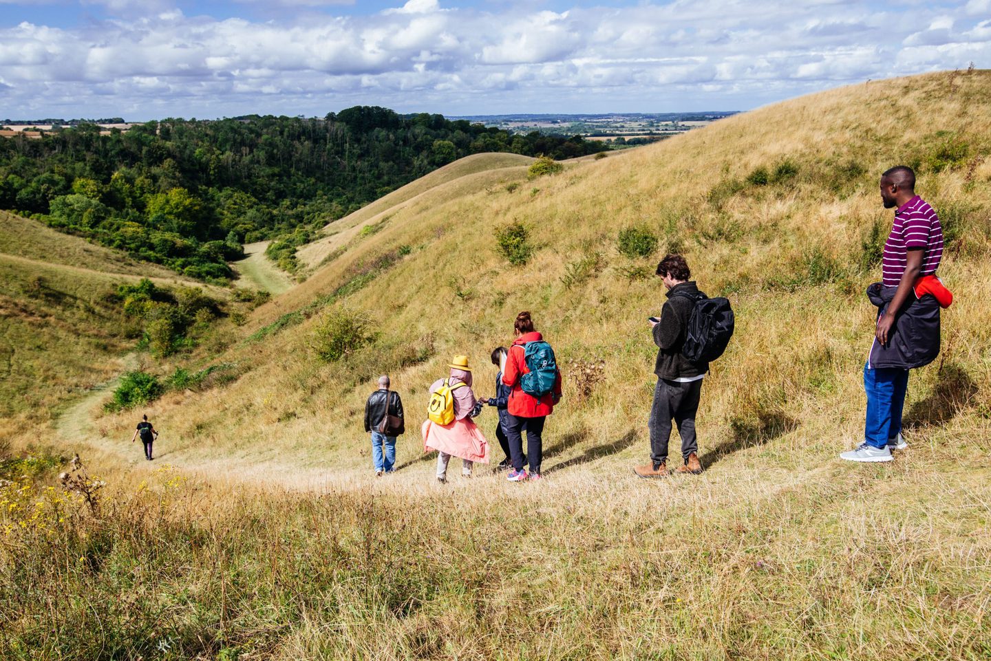 A group of seven people hiking down a grassy hill in a scenic, hilly landscape. The hikers are wearing various outdoor clothing and backpacks. The terrain is covered with dry grass and scattered bushes, with lush green trees visible in the distance under a partly cloudy sky.