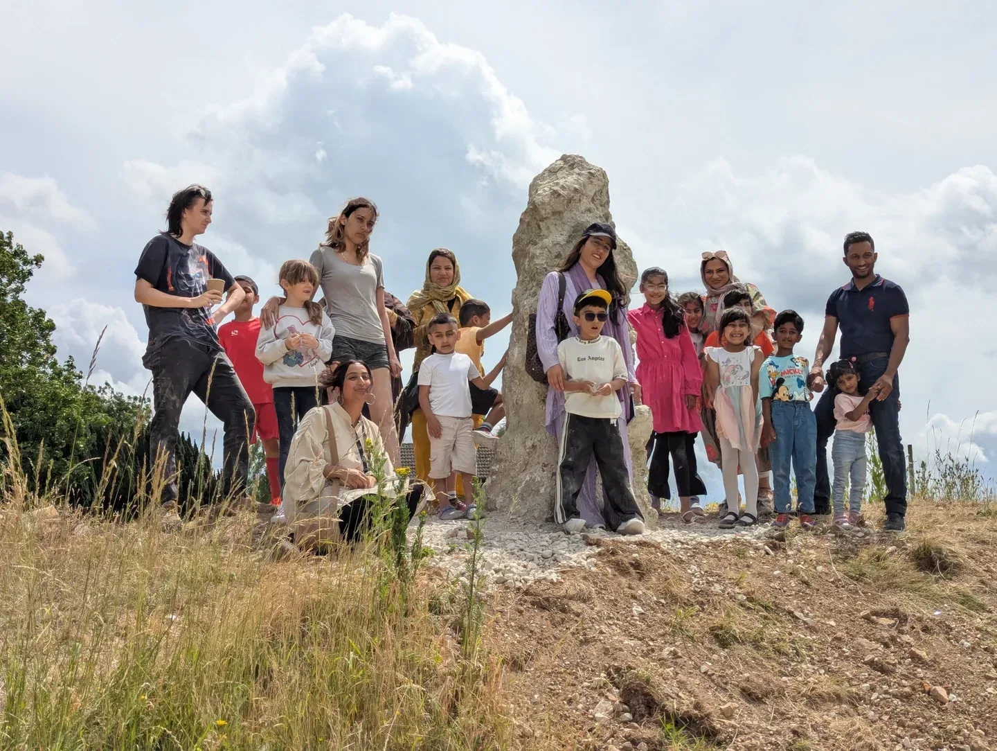 A group of people, including adults and children, gathered around a large standing stone on a grassy hill. The sky is partly cloudy, and the setting is outdoors in nature. Some individuals are standing while others are crouching or sitting on the ground.