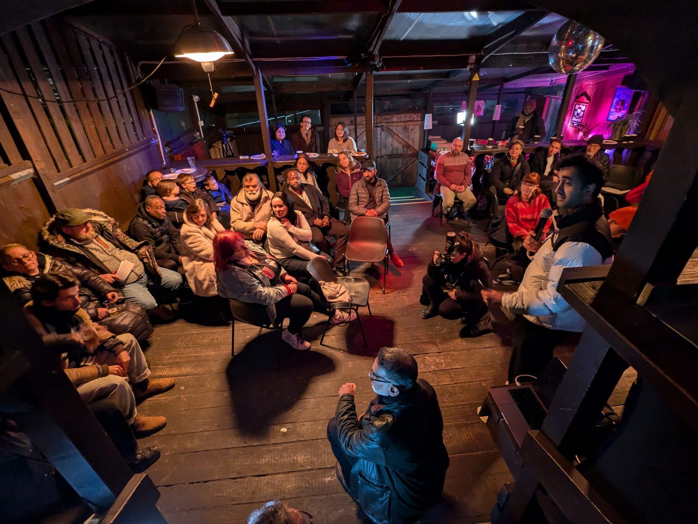 A group of people seated in a semi-circle inside a dimly lit room with wooden floors and walls. Some individuals are sitting on chairs while others are standing or sitting on the floor. The room has a cozy, rustic atmosphere with various decorations and lights, including a disco ball hanging from the ceiling. The focus appears to be on a central figure who is speaking or presenting to the group.