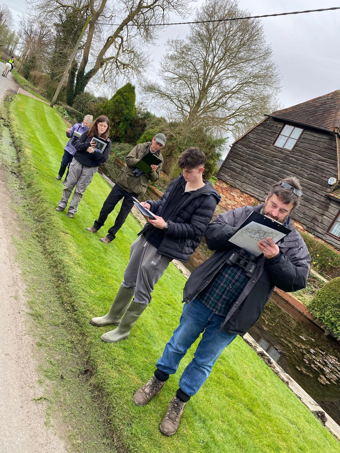 A group of six people are standing on a grassy area next to a narrow road, holding notebooks and writing or drawing. They are dressed in outdoor clothing, including jackets and boots. In the background, there is a wooden house with a brick foundation and leafless trees. The scene appears to be in a rural setting with overcast weather.